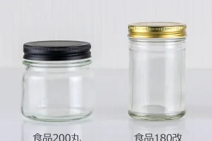 黒いキャップの短いガラス瓶（食品200丸）と、金色キャップの細長いガラス瓶（食品180改）が並んで置かれている。どちらも透明で、背景は淡い縦ラインの壁